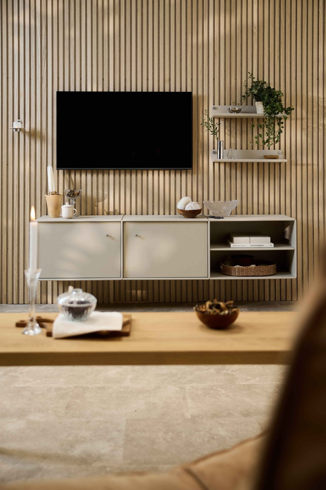 Salón moderno con el mueble de televisión «Arka» de WoodUpp, sofás modulares, estanterías de estilo nórdico con plantas y elementos decorativos, y una vela sobre la mesita de centro.